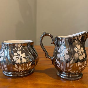 Peut inclure: Un ensemble de crémier et sucrier argentés. L'ensemble présente un motif floral blanc. Le crémier a une anse et un bec verseur. Le sucrier est rond avec un motif floral assorti. Les deux pièces ont une finition brillante et réfléchissante.