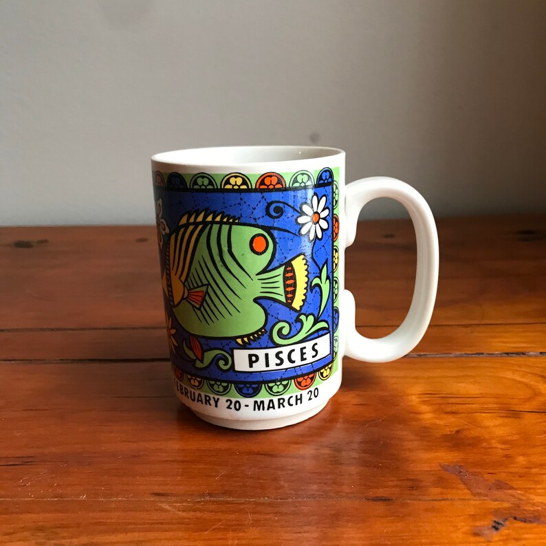May include: White ceramic mug with a blue, green, yellow, and orange design featuring a stylized fish and the text "Pisces February 20 - March 20".