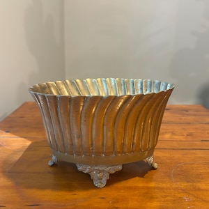 May include: An oval brass bowl featuring a vertical fluted or ribbed pattern along the exterior walls. The bowl is supported by three ornate, decorative feet with intricate detailing, and the metal surface displays a warm, metallic gold tone.
