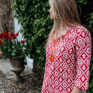 May include: A red and white patterned tunic dress with three-quarter sleeves. The dress has a geometric design and orange tassel detailing at the neckline. The model is standing outside near a green bush and red flowers.