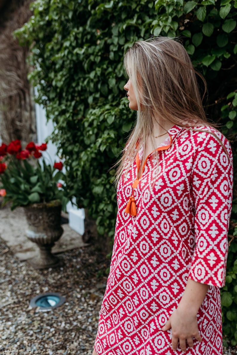 May include: A red and white patterned tunic dress with three-quarter sleeves. The dress has a geometric design and orange tassel detailing at the neckline. The model is standing outside near a green bush and red flowers.