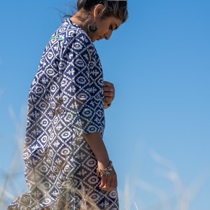 May include: A woman in a long, navy blue and white patterned dress with three-quarter sleeves. The dress features a geometric design. She wears turquoise earrings and a gold bracelet. The background is a clear blue sky and tall grass.