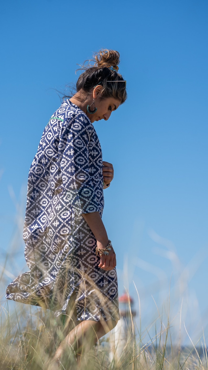 May include: A woman in a long, navy blue and white patterned dress with three-quarter sleeves. The dress features a geometric design. She wears turquoise earrings and a gold bracelet. The background is a clear blue sky and tall grass.