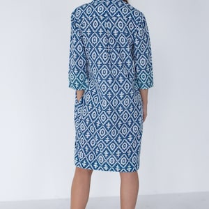 May include: A blue and white patterned dress, knee-length with three-quarter sleeves. The dress has a geometric design and a collared neckline. The model is wearing white sandals. The dress has pockets.