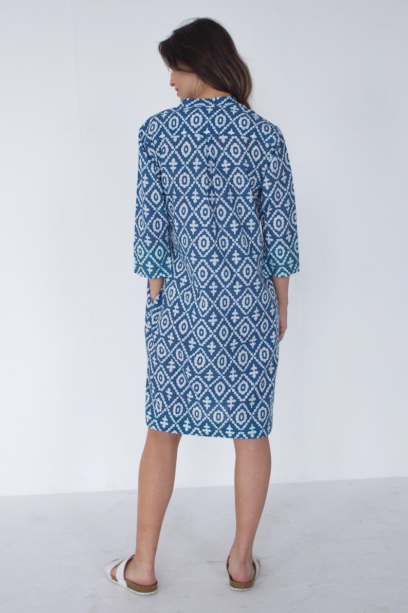 May include: A blue and white patterned dress, knee-length with three-quarter sleeves. The dress has a geometric design and a collared neckline. The model is wearing white sandals. The dress has pockets.