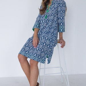 May include: A blue and white patterned dress with a V-neck and three-quarter sleeves. The dress has a geometric design and green trim. The model is wearing white sandals and is sitting on a white stool.
