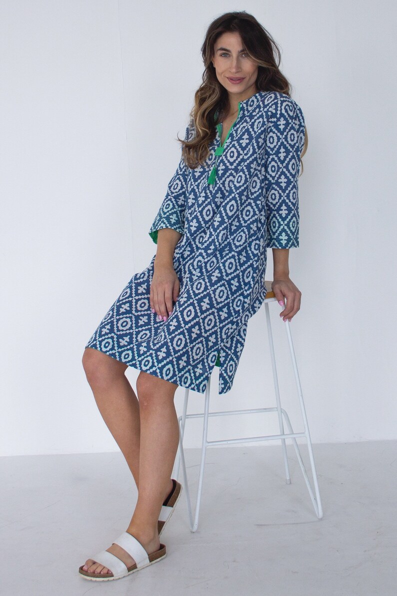 May include: A blue and white patterned dress with a V-neck and three-quarter sleeves. The dress has a geometric design and green trim. The model is wearing white sandals and is sitting on a white stool.