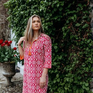 May include: A red and white patterned dress with a mandarin collar and three-quarter sleeves. The dress has an orange tassel and orange trim. The background features green foliage and a stone wall. The dress is knee-length.
