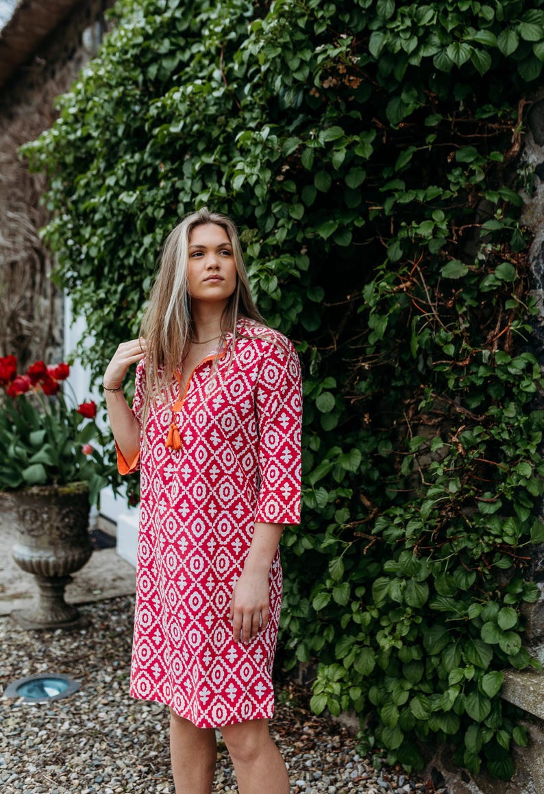 May include: A red and white patterned dress with a mandarin collar and three-quarter sleeves. The dress has an orange tassel and orange trim. The background features green foliage and a stone wall. The dress is knee-length.
