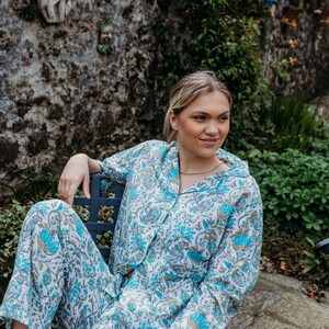 May include: A light blue and white floral print pajama set. The long-sleeved button-down shirt and matching trousers feature a teal and tan floral pattern. The person is sitting on a dark blue chair.