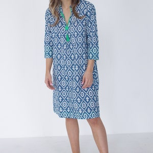 May include: A blue and white patterned knee-length dress with three-quarter sleeves and a V-neck with a green tassel. The dress has green trim. The person is wearing white sandals.