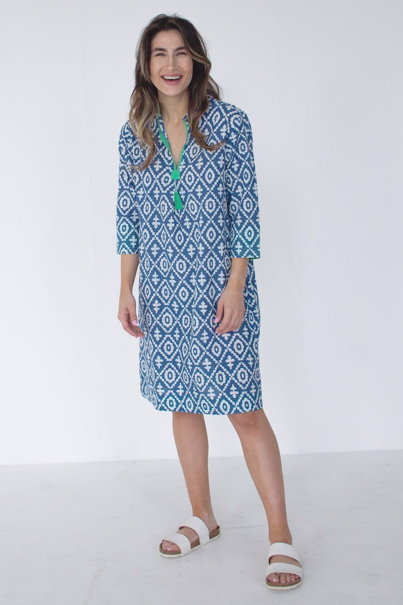 May include: A blue and white patterned knee-length dress with three-quarter sleeves and a V-neck with a green tassel. The dress has green trim. The person is wearing white sandals.