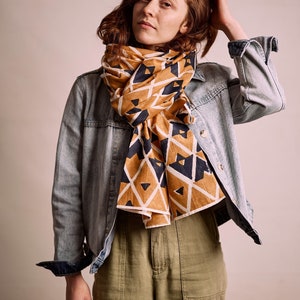 K&ouml;nnte beinhalten: Ein gemusterter Schal in Senfgelb und Marineblau, drapiert &uuml;ber einer Jeansjacke. Der Schal hat ein geometrisches Design aus Dreiecken und Rauten. Das Model tr&auml;gt eine olivgr&uuml;ne Hose.