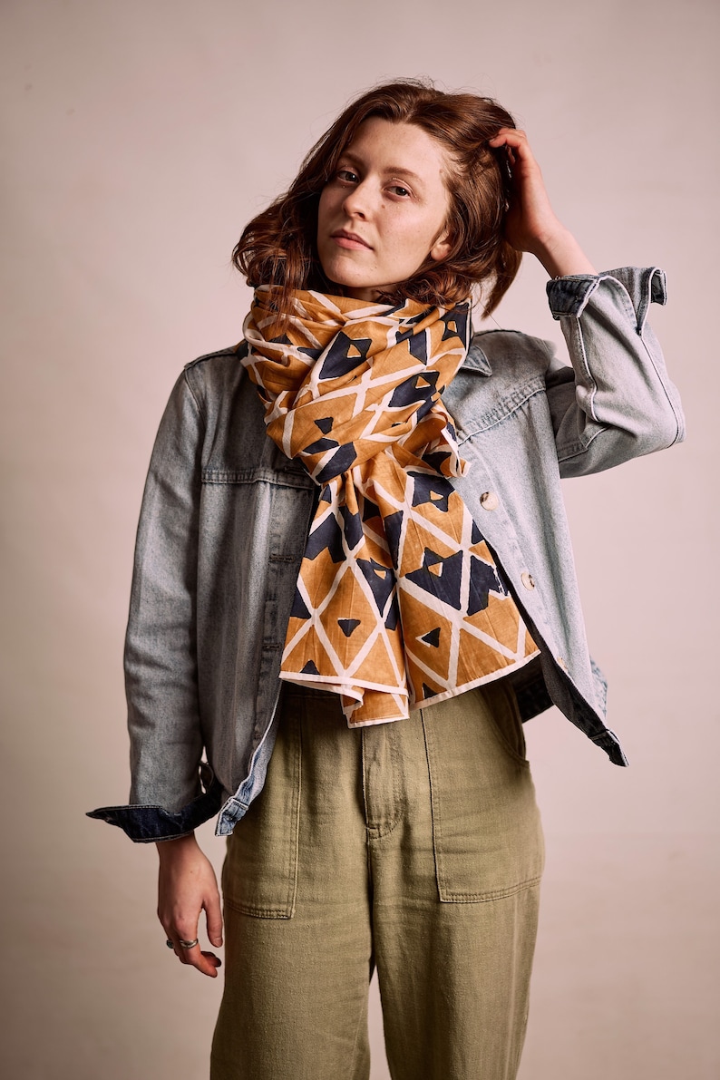 K&ouml;nnte beinhalten: Ein gemusterter Schal in Senfgelb und Marineblau, drapiert &uuml;ber einer Jeansjacke. Der Schal hat ein geometrisches Design aus Dreiecken und Rauten. Das Model tr&auml;gt eine olivgr&uuml;ne Hose.