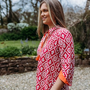 May include: A red and white patterned tunic dress with orange trim and tassels. The dress has a geometric design and a mandarin collar. The sleeves are three-quarter length. The model is smiling outdoors.