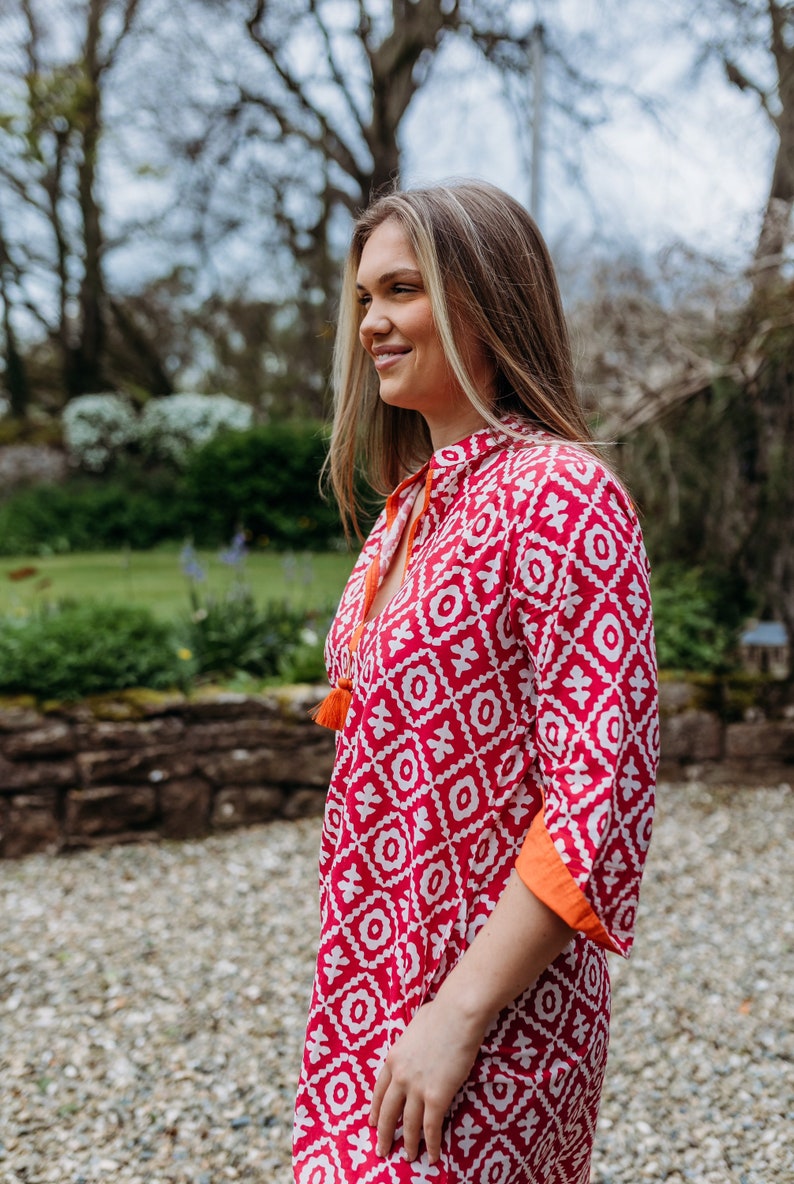 May include: A red and white patterned tunic dress with orange trim and tassels. The dress has a geometric design and a mandarin collar. The sleeves are three-quarter length. The model is smiling outdoors.