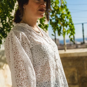 K&ouml;nnte beinhalten: Eine Frau tr&auml;gt eine wei&szlig;e Blusenbluse mit einem zarten Blumenmuster. Die Bluse hat einen R&uuml;schenkragen und Dreiviertel&auml;rmel. Die Frau steht vor einer gr&uuml;nen, belaubten Pflanze.