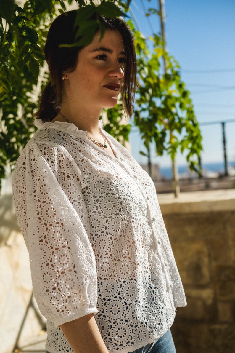 K&ouml;nnte beinhalten: Eine Frau tr&auml;gt eine wei&szlig;e Blusenbluse mit einem zarten Blumenmuster. Die Bluse hat einen R&uuml;schenkragen und Dreiviertel&auml;rmel. Die Frau steht vor einer gr&uuml;nen, belaubten Pflanze.