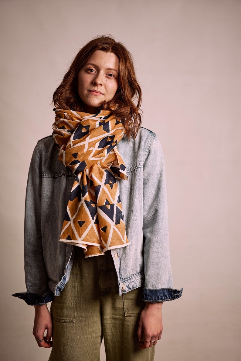 K&ouml;nnte beinhalten: Senffarbener Schal mit einem geometrischen Muster aus wei&szlig;en und marineblauen Dreiecken. Der Schal ist um Hals und Schultern drapiert. Die Person tr&auml;gt eine Jeansjacke und eine olivgr&uuml;ne Hose.