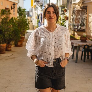 K&ouml;nnte beinhalten: Eine Frau tr&auml;gt eine wei&szlig;e H&auml;kelbluse mit einem zarten Blumenmuster und schwarze Jeans-Shorts. Sie steht in einer engen Gasse mit wei&szlig;en Geb&auml;uden auf beiden Seiten.