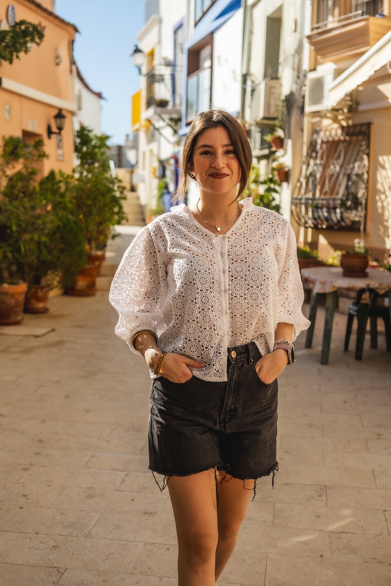K&ouml;nnte beinhalten: Eine Frau tr&auml;gt eine wei&szlig;e H&auml;kelbluse mit einem zarten Blumenmuster und schwarze Jeans-Shorts. Sie steht in einer engen Gasse mit wei&szlig;en Geb&auml;uden auf beiden Seiten.