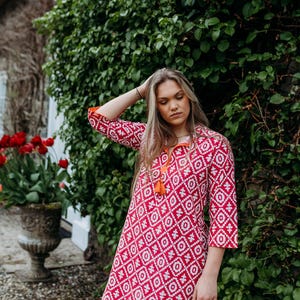 May include: A red and white patterned dress with three-quarter sleeves and an orange tassel neckline. The dress has a geometric design and is worn outdoors near a green hedge and red tulips.