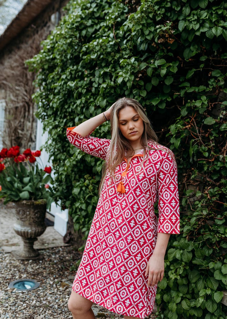 May include: A red and white patterned dress with three-quarter sleeves and an orange tassel neckline. The dress has a geometric design and is worn outdoors near a green hedge and red tulips.