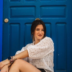 K&ouml;nnte beinhalten: Eine Frau in einem wei&szlig;en Spitzenoberteil und schwarzen Jeans-Shorts sitzt vor einer leuchtend blauen T&uuml;r. Sie l&auml;chelt in die Kamera.