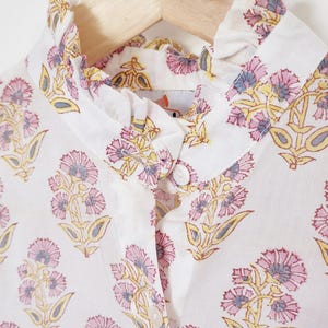 May include: White button-up shirt with a high ruffled collar and a floral pattern. The pattern features pink and yellow flowers with blue accents. The shirt is hanging on a wooden hanger.
