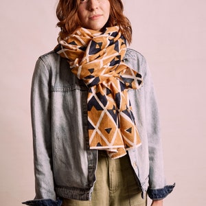 K&ouml;nnte beinhalten: Ein senfgelber und marineblauer Schal mit Muster ist um den Hals gelegt. Der Schal wird mit einer hellblauen Jeansjacke und einer olivgr&uuml;nen Hose kombiniert. Das geometrische Design des Schals setzt einen Farbakzent.