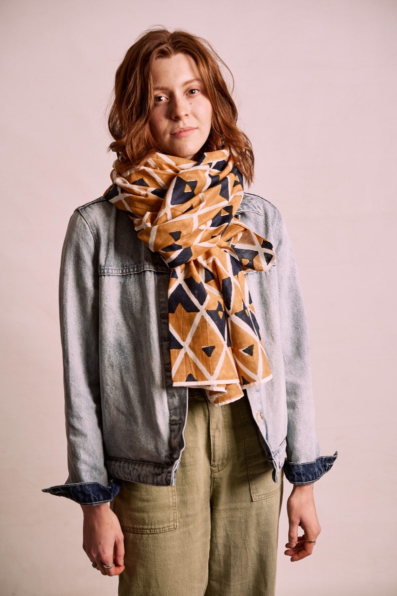 K&ouml;nnte beinhalten: Ein senfgelber und marineblauer Schal mit Muster ist um den Hals gelegt. Der Schal wird mit einer hellblauen Jeansjacke und einer olivgr&uuml;nen Hose kombiniert. Das geometrische Design des Schals setzt einen Farbakzent.