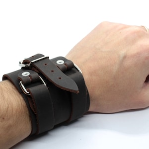 May include: A brown leather cuff bracelet with a silver buckle and two straps. The bracelet is adjustable and can be worn on either wrist.