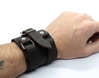 May include: A brown leather cuff bracelet with a silver buckle and two straps. The bracelet is adjustable and can be worn on either wrist.