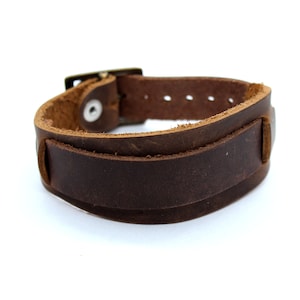 Könnte beinhalten: Ein braunes Lederarmband mit einer Schließe. Das Armband ist aus dickem, strapazierfähigem Leder gefertigt und hat ein schlichtes, klassisches Design.