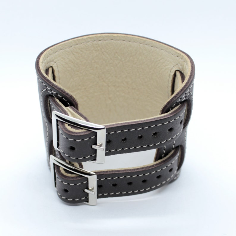 May include: A brown leather wrist strap with two adjustable buckles. The strap is stitched with white thread and has a cream-colored lining.