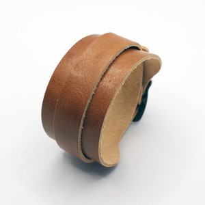 Puede incluir: Un brazalete de cuero marrón con cierre de hebilla. El brazalete está hecho de dos capas de cuero, y la hebilla es de metal.