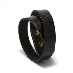 May include: A dark brown leather wrap bracelet with brass-colored screw details. The bracelet is designed to wrap around the wrist, showcasing a minimalist and classic style. The leather appears to be smooth and well-crafted.