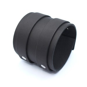 May include: A black leather cuff bracelet with silver metal accents. The bracelet is made of multiple layers of leather and has a simple, minimalist design.