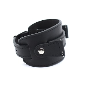 Pode incluir: Uma pulseira de relógio de couro preto com uma fivela prateada e um fecho de botão único.
