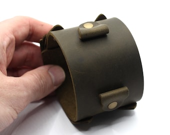 May include: A wide, olive green leather cuff bracelet with brass-colored button closures. The bracelet is thick and has a slightly distressed appearance. The cuff is held by a hand against a white background.