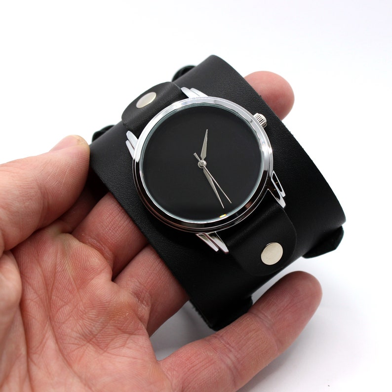 May include: A black leather cuff watch with a silver face and black hands. The watch is on a person's wrist.