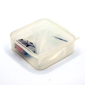 May include: A clear plastic container with a silver watch movement, a small battery, and a few small metal parts inside.