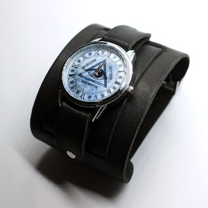 May include: A silver wristwatch with a black leather cuff band. The watch face has a white background with a black and white all-seeing eye design and Roman numeral hour markers.