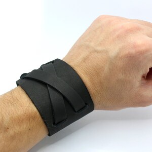 May include: A black leather bracelet with a braided design. The bracelet is worn on a person's wrist.