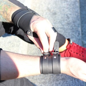 May include: A black leather wrist cuff with a silver buckle and two silver studs. The cuff is adjustable and has a layered design.