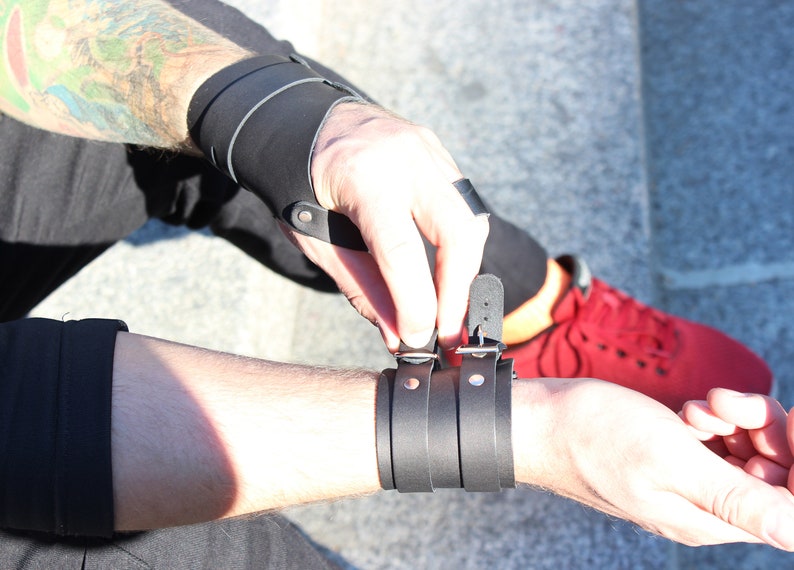 May include: A black leather wrist cuff with a silver buckle and two silver studs. The cuff is adjustable and has a layered design.
