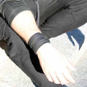 May include: A black leather bracelet with three straps. The bracelet is worn on the wrist of a person wearing black pants.