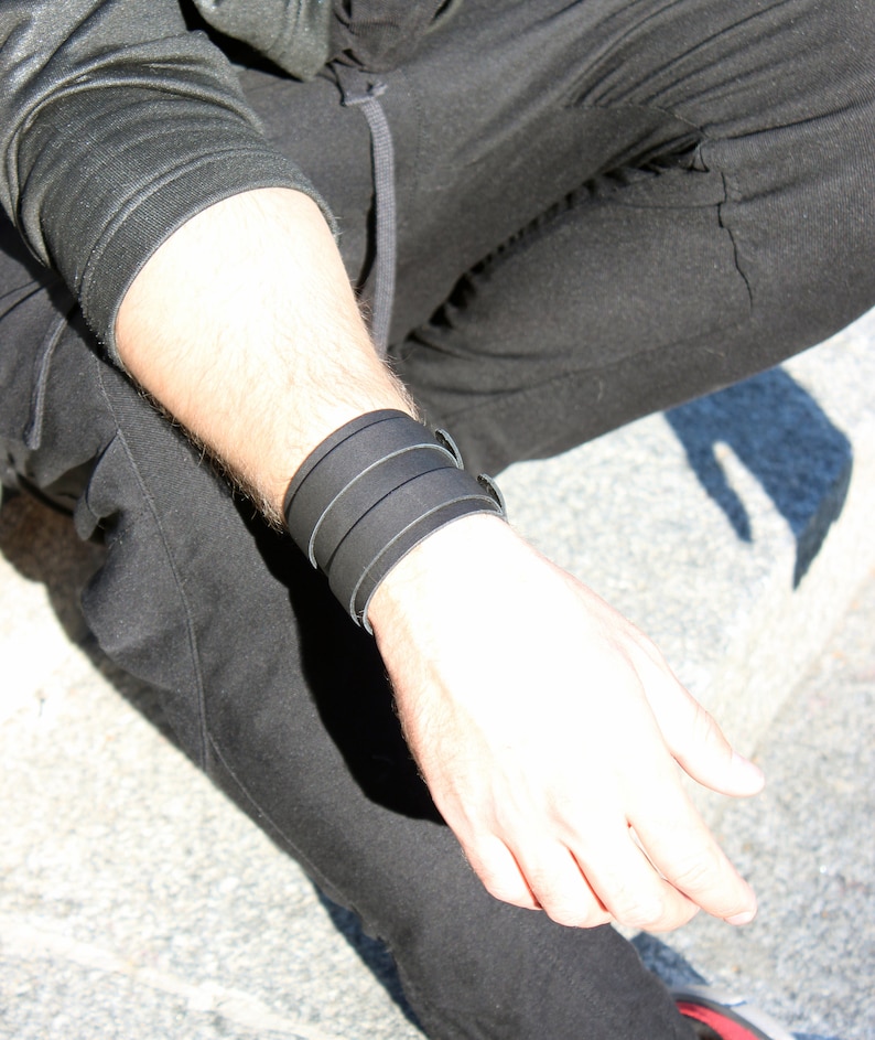 May include: A black leather bracelet with three straps. The bracelet is worn on the wrist of a person wearing black pants.