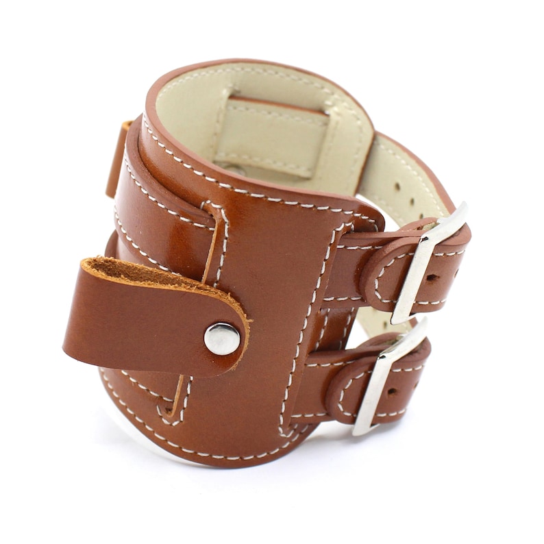 May include: A brown leather watch band with two adjustable straps and a buckle closure. The band has white stitching and a metal button.