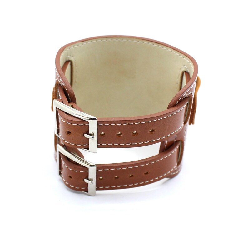 May include: A brown leather watch band with two silver buckles. The band has white stitching and a smooth leather interior.